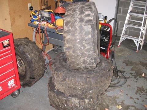 1995 GMC Sierra 1500 - A stack of four large, knobby off-road tires are piled in a garage next to a red toolbox and a workbench.