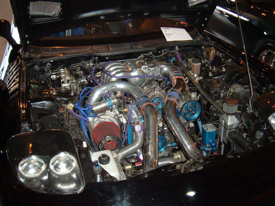 Black car engine bay with twin turbochargers, chrome piping, and blue accents. A white paper is visible on top