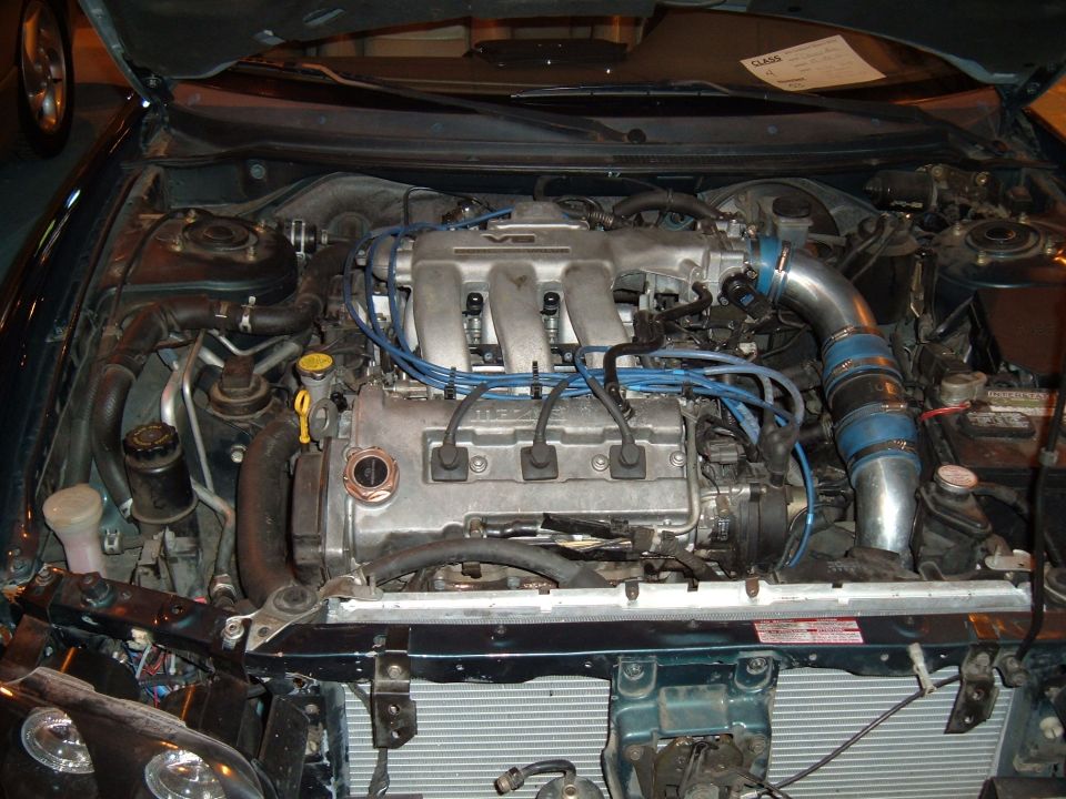 Engine bay of a 1991 Ford Probe, V6 engine with blue spark plug wires, silver intake piping, and a white label on the hood