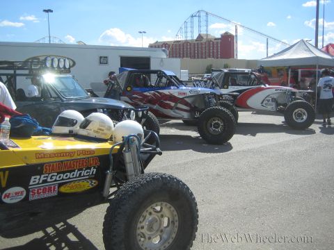 1990 Ford Bronco - row of open-cockpit off-road race buggies and trucks, including a yellow car with helmets, parked at an event with a roller coaster behind.