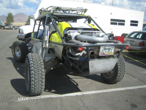 1990 Ford Bronco - rear of a yellow-green off-road race buggy with twin upturned exhaust pipes, tubular cage and a license plate, parked beside trailers.