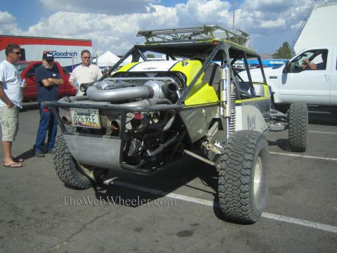 1990 Ford Bronco - front of a yellow and bare-metal off-road race buggy with twin turbochargers and a light bar, with onlookers standing beside it in a lot.
