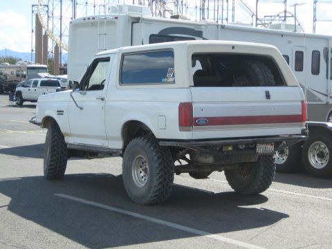 1990 Ford Bronco - rear three-quarter view of a white lifted Ford Bronco on large mud tires, with a red trim stripe, parked in a lot near RVs.