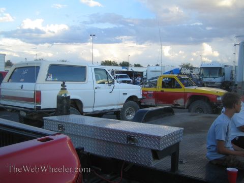 1990 Ford Bronco - pit area at dusk with a white Bronco, a yellow and red race truck and trailers, a diamond-plate toolbox in front and a seated boy at right.
