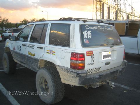 1990 Ford Bronco - mud-splattered white Jeep Grand Cherokee with lift kit and Desert Dog Racing graphics, number 1615, parked in a lot under a sunset sky.