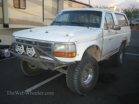 1990 Ford Bronco - white lifted Ford Bronco with a tubular front bumper, twin Hella driving lights and muddy fender flares parked beside an RV.