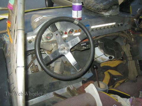 1990 Ford Bronco - stripped interior of an off-road race car showing a four-spoke steering wheel, gauge cluster, switch panel and exposed wiring.