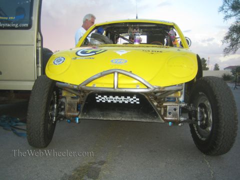 1990 Ford Bronco - yellow off-road race buggy photographed head-on, revealing its tubular front bumper, A-arm suspension and checkered decals.