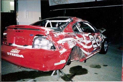 1995 Ford Mustang - A damaged 1995 Ford Mustang race car sits on stands, showing extensive bodywork and racing livery.