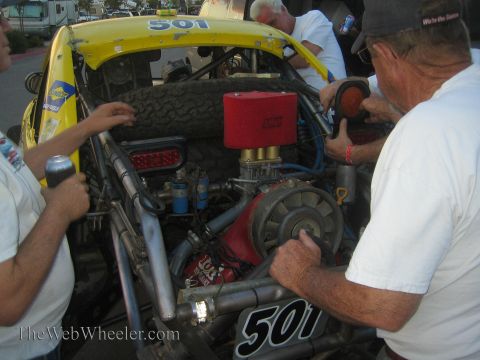 1990 Ford Bronco - Crew members in white shirts work on the rear-mounted engine of a yellow number 501 off-road race car, with a red air filter visible.