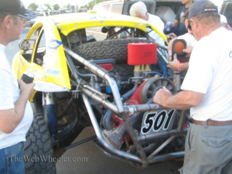 1990 Ford Bronco - Two crew members service the exposed tubular rear chassis and engine of a yellow number 501 off-road race car in a paddock.