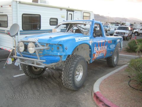 1990 Ford Bronco - blue Horizon Realty off-road race truck, number 743, with tubular front bumper and driving lights parked on pavement beside RVs.