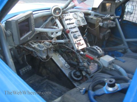 1990 Ford Bronco - cockpit of a blue off-road race car showing gauges, a switch panel, a GPS-style screen, removable steering wheel and exposed wiring.