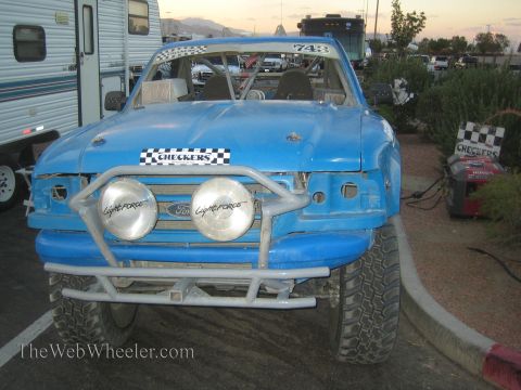 1990 Ford Bronco - blue off-road race truck, number 743, with Checkers and Ford decals, tubular front bumper and twin Lightforce driving lights at dusk.
