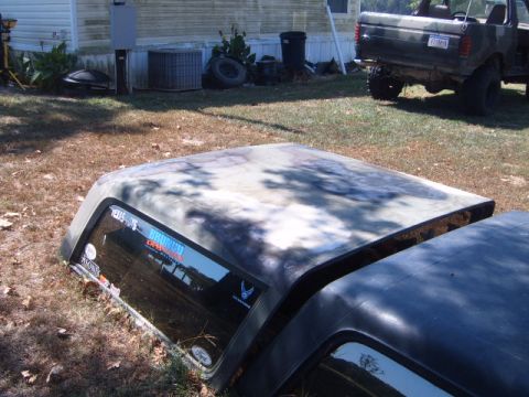 1988 Ford Bronco - A camouflaged Bronco hardtop lies on the ground next to another vehicle part. The hardtop has several stickers on its window.