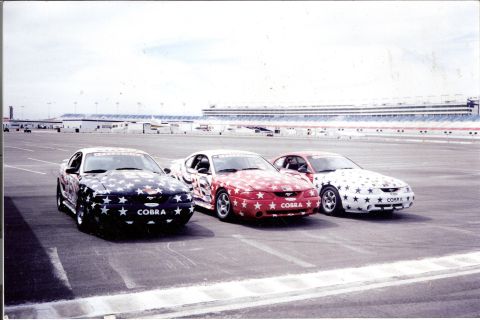 1995 Ford Mustang - Three star-spangled Ford Mustangs are lined up on a racetrack, with grandstands in the background.