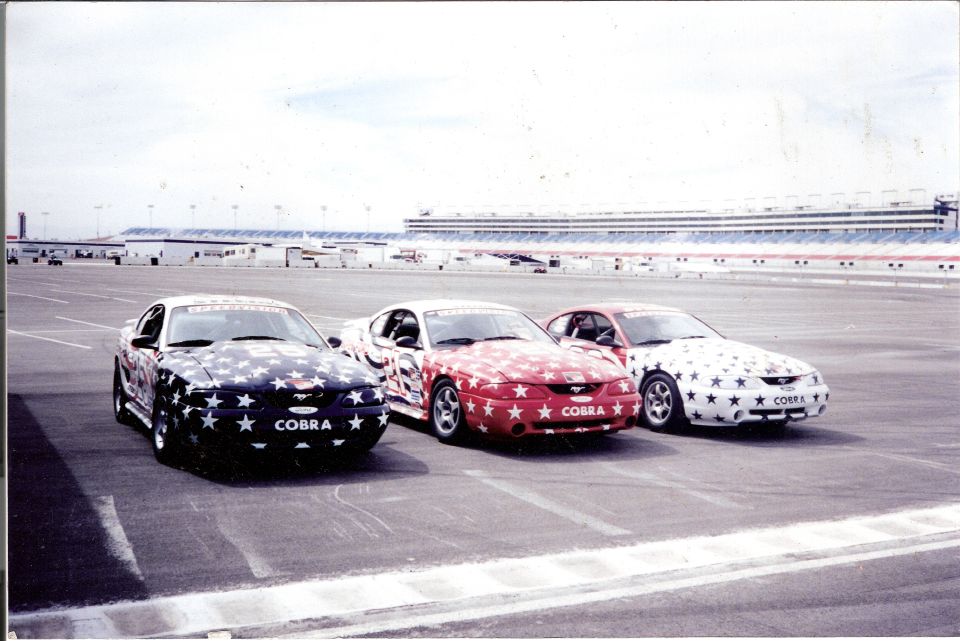 Three Ford Mustangs, two red and white, one black and white, all with star graphics and 'COBRA' on the front. They are parked on a racetrack