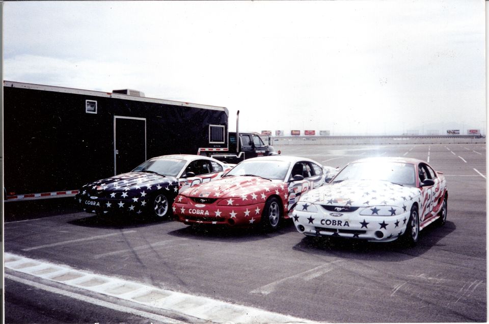 Three 1995 Ford Mustangs with star and stripe paint jobs parked on asphalt. The middle car has a red base with white stars, the left car has a dark base with white stars, and the right car has a white base with red and blue stars and stripes. All have ...