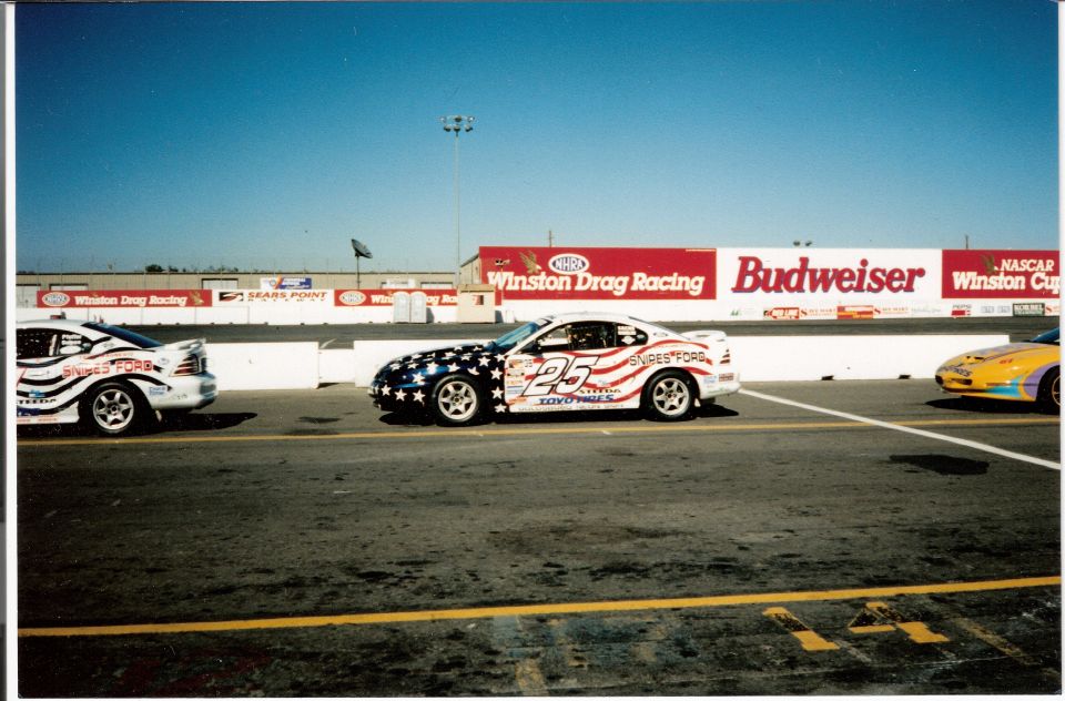 Two race cars, one with stars and stripes livery and number 25, on a drag strip with sponsor banners in the background