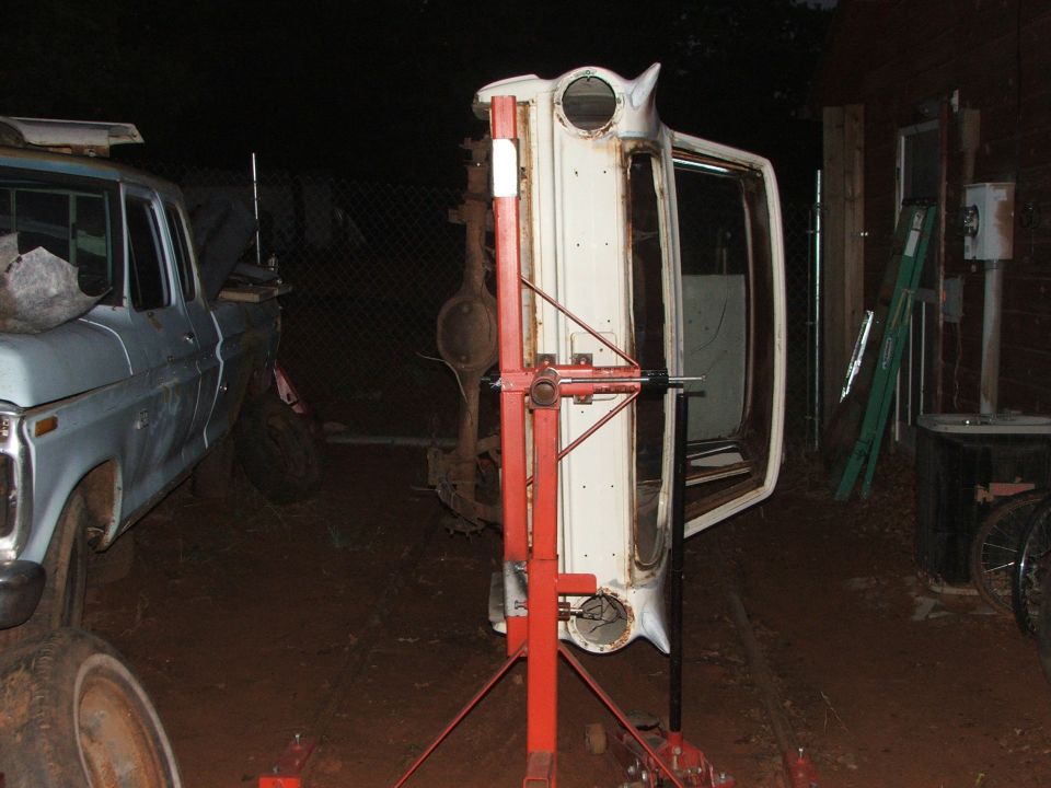 White car body upside down on a red stand, with a light blue pickup truck to the left