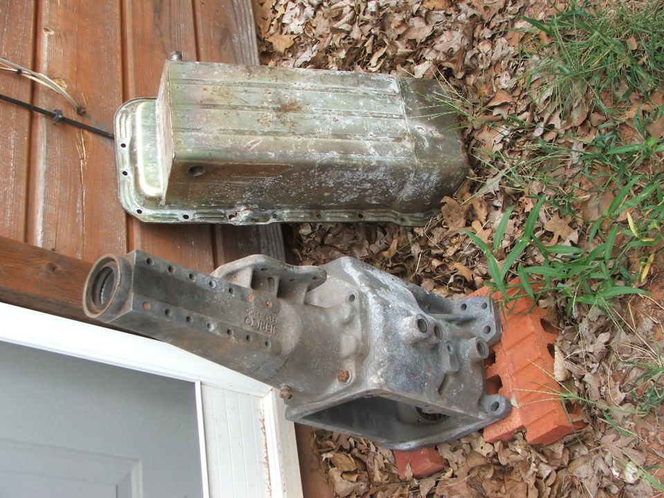 Car transmission and oil pan sitting on the ground next to a wooden wall