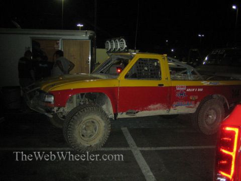 1990 Ford Bronco - red and yellow off-road race truck, number 744, with a roof-mounted light pod parked at night in a paddock area.