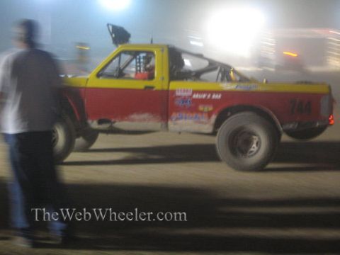 1990 Ford Bronco - red and yellow off-road race truck, number 744, captured in motion blur on a dirt course at night with a bystander watching.