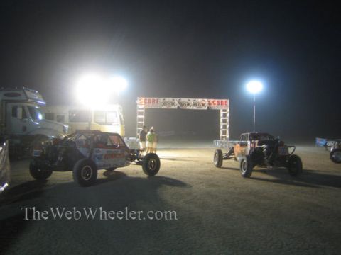 1990 Ford Bronco - Two off-road race buggies idle on a dirt staging area near a brightly lit SCORE start arch in the desert at night.