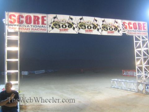 1990 Ford Bronco - SCORE International off-road racing start arch lit at night, hung with event banners over a dirt course, a man standing at left.