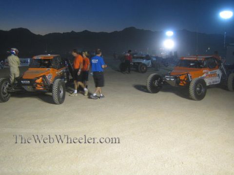 1990 Ford Bronco - Orange off-road race buggies and crew members gather on a dirt staging area at dusk, mountains and floodlights in the background.