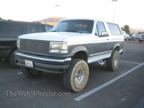 1990 Ford Bronco - silver lifted full-size Ford Bronco with a billet grille and oversized tires parked in a lot, mountains behind at dusk.