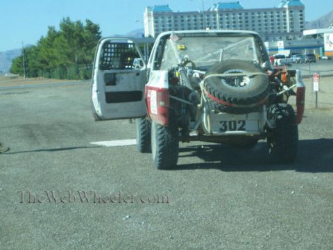 1990 Ford Bronco - rear of a white off-road race truck, number 302, with a bed-mounted spare tire and an open door, parked on gravel near a casino.