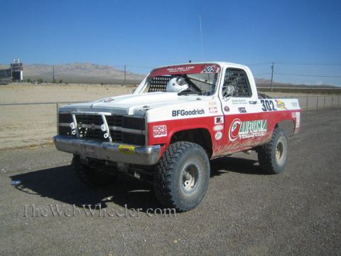 1990 Ford Bronco - red and white lifted Chevrolet pickup race truck numbered 302 with BFGoodrich decals sits on dirt in a desert pit area.