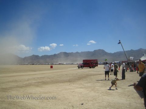 1990 Ford Bronco - Spectators and a boom mic watch across a dusty open desert flat with a distant red support truck and mountains behind.
