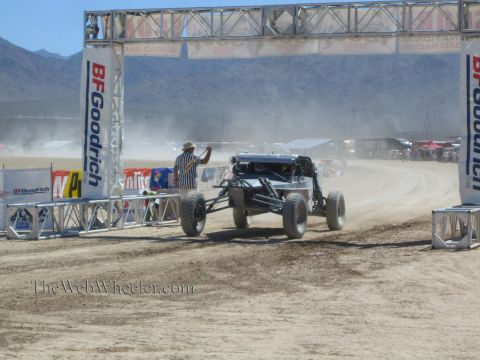 1990 Ford Bronco - black off-road race buggy passes under a BFGoodrich start-finish arch on a dusty desert course as an official signals.
