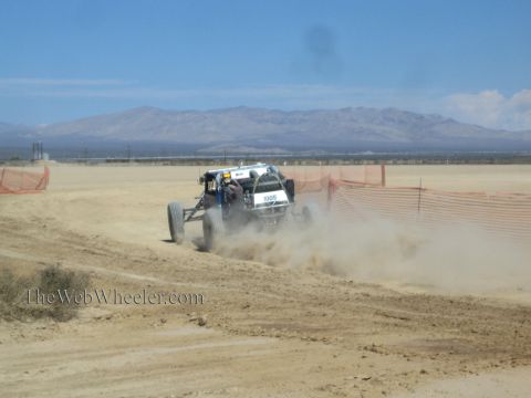 1990 Ford Bronco - white number 1005 off-road race buggy throws up a cloud of dust rounding a fenced bend on a desert course.