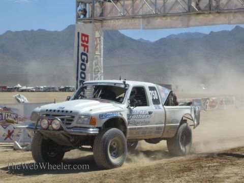 1990 Ford Bronco - white number 758 off-road race pickup with BFGoodrich decals races through desert dust under a banner-lined arch.