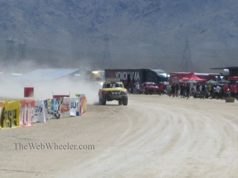 1990 Ford Bronco - off-road race truck raises a cloud of dust on a desert course lined with vendor banners, semis, and spectators.