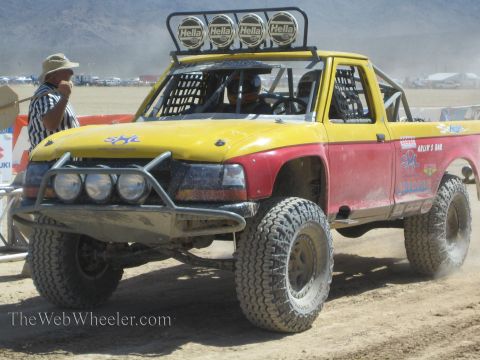 1990 Ford Bronco - yellow and red off-road race truck fitted with four Hella roof lights waits on a desert course as an official stands by.