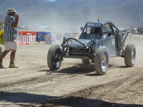 1990 Ford Bronco - black number 1616 tube-frame off-road race buggy sits on a desert course as a striped-shirt official gestures nearby.