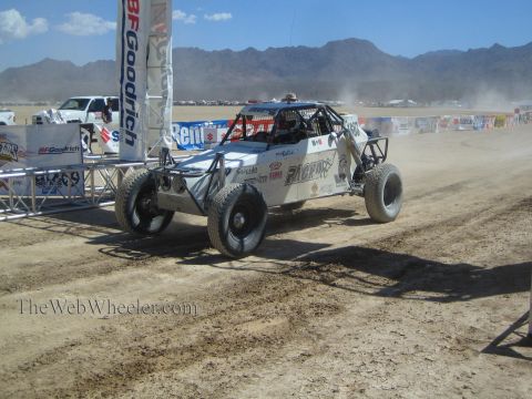 1990 Ford Bronco - white off-road race buggy races past BFGoodrich and sponsor banners on a desert course with mountains behind.