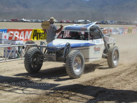 1990 Ford Bronco - blue and white off-road race buggy waits at the start line as a striped-shirt official signals past FRAM and Suzuki banners.