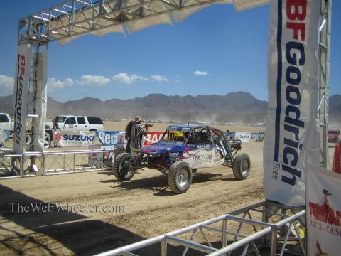 1990 Ford Bronco - off-road race buggy passes under a BFGoodrich start arch as a photographer crouches nearby on the dusty desert course.