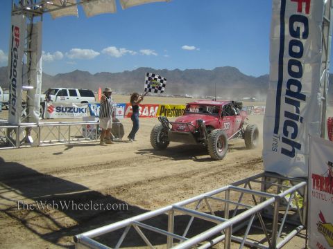 1990 Ford Bronco - red number 302 off-road race truck takes the checkered flag waved by a woman under a BFGoodrich arch in the desert.