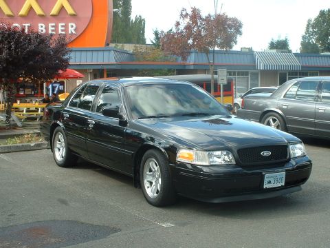 2000 Ford Crown Victoria - A black Ford Crown Victoria is parked in a lot, with a building and another car visible in the background.