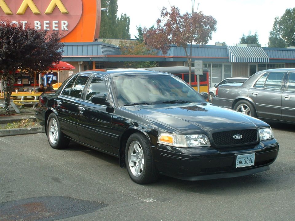 Black Ford Crown Victoria parked in front of a building with a large orange sign. Another gray car is parked nearby