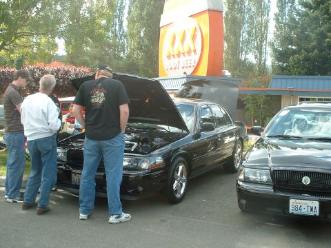 2000 Ford Crown Victoria - Two black sedans are displayed at an outdoor event, with one car's hood open for viewing.
