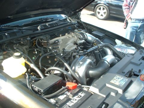 2000 Ford Crown Victoria - The engine bay of a car, a prominent supercharger and various hoses and components. A second vehicle is partially visible.