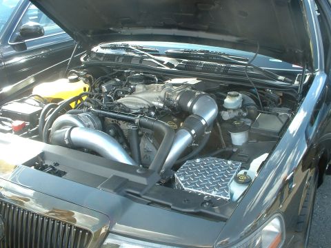 2000 Ford Crown Victoria - The engine bay of a car features a supercharger and custom intake piping, with another vehicle visible in the background.