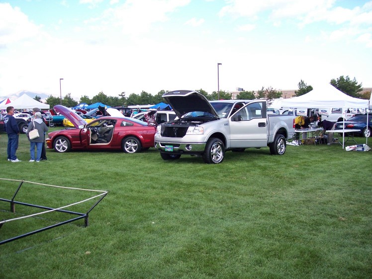 Silver pickup truck with hood open next to a red coupe with door open, parked on grass at an outdoor event